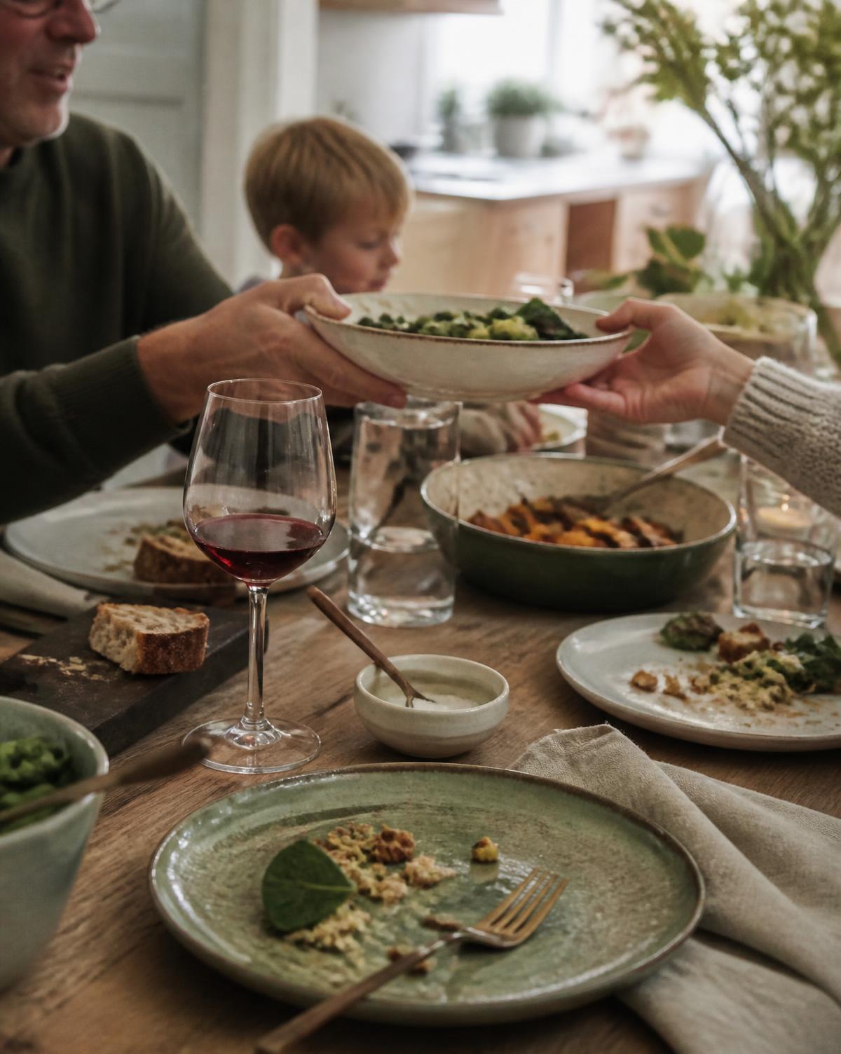 A bowl of food passed between hands across a wooden family dining table at home, with stoneware plates and a glass of wine